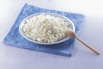 Cottage cheese on blue napkin. Cheese on white plate with wooden spoon. Soft cheese for diet. Healthy dairy product for breakfast