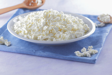 Cottage cheese on blue napkin. Cheese on white plate with wooden spoon. Soft cheese for diet. Healthy dairy product for breakfast