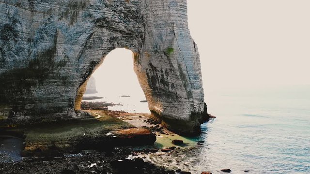 Drone flying right along epic natural rock arch at famous white alabaster seaside cliffs at Etretat Normandy on sunset.