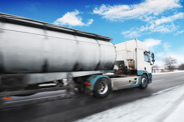 Fototapeta na wymiar Big metal fuel tanker truck in motion shipping fuel on the winter countryside road with snow against blue sky