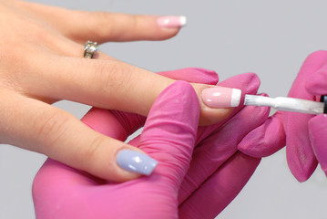 Obraz na płótnie Canvas A beautiful female hand with a manicure in two colors - blue and pink - lies on the hand of the manicurist in a pink hygienic glove. The master applies white varnish on the tip of the pink nail, produ