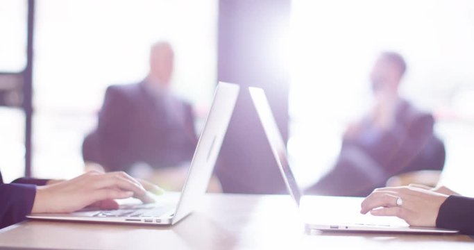 4K Businesswomen using laptops in bright modern office with businessmen blurred in background