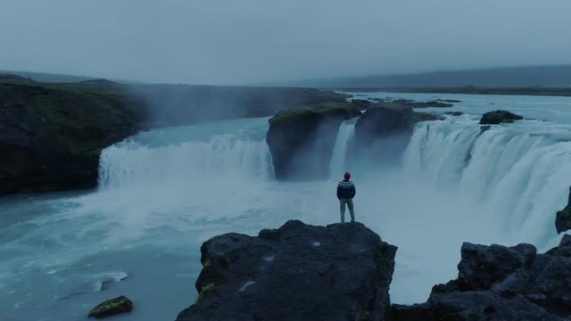 Epic adventure seeker drone shot of urban nomad traveller,hipster young millennial man stand on edge of cliff near waterfall in iceland, confident and brave, search for new exciting horizons and ideas