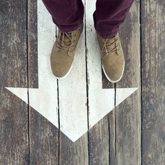 White arrow sign on wooden floor. Man's legs standing on arrow sign painted on the floor. Direction...