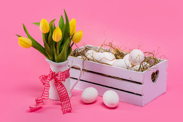 Easter. Eggs in a white wooden box with hay and yellow tulips in a white jug. Pink background.