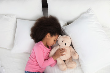 Cute little African-American girl with teddy bear sleeping in bed, top view