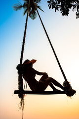 Silhouette woman wear bikini and straw hat swing the swings at the beach on summer vacation at sunset. Girl in swimwear sit on swings and watch beautiful sunset. Summer vibes. Woman travel alone