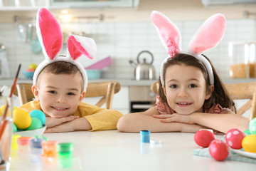 Cute children with bunny ears headbands and painted Easter eggs sitting at table in kitchen