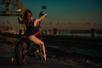 Red-haired woman in sexy lingerie in high heels sits on a motorcycle. Attractive red-haired girl sits on a motorcycle at sunset on the background of the industrial landscape