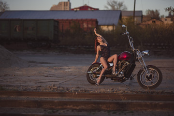 Red-haired woman in sexy lingerie in high heels sits on a motorcycle. Attractive red-haired girl sits on a motorcycle at sunset on the background of the industrial landscape