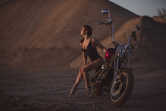 Red-haired woman in sexy lingerie in high heels sits on a motorcycle. Attractive red-haired girl sits on a motorcycle at sunset on the background of the industrial landscape