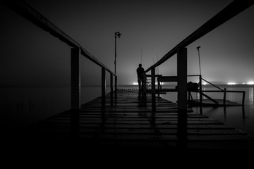Silhouette of a man standing on a wooden dock in the dark staring at the emptiness of a lake with glowing lights on the opposite side
