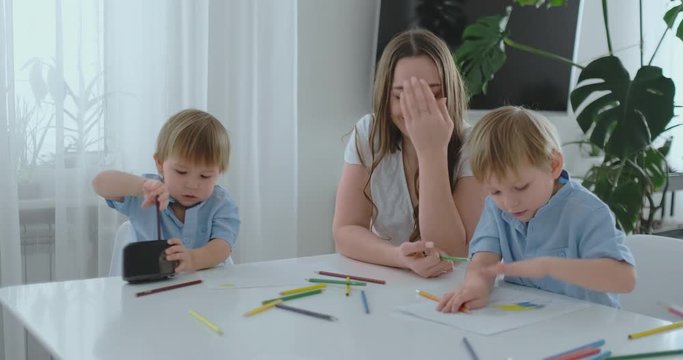 Mom sitting at the table with two sons Boy sharpening a colored pencil in a sharpener sitting at the kitchen table. The second boy draws a picture with pencils