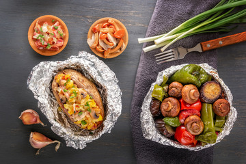 Baked potatoes with bacon, onions and baked vegetables in foil - tomatoes, eggplants, peppers on a gray wooden table. Top view