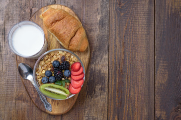 granola with berries and fruit, croissant, glass of milk on wooden background