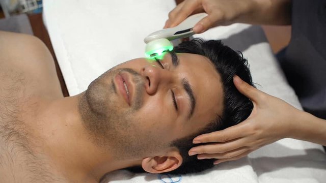 Medium shot of Asian man lying on spa bed and having facial treatment with laser