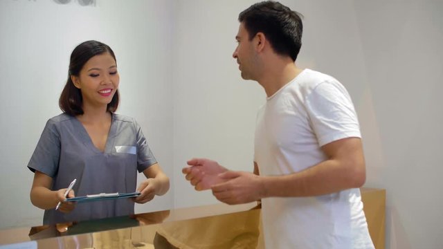 Medium shot of Asian receptionist of spa center greeting male client and giving him form for filling in