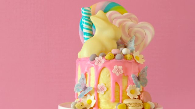On trend Easter candy land drip cake decorated with lollipops, cand eggs and white chocolate bunny in pink party table setting.