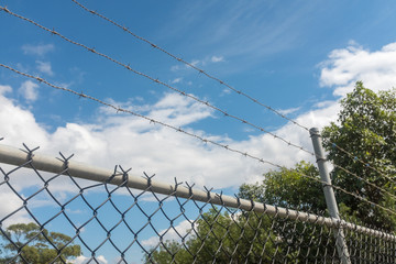 Close up of barbed wire and metal fence against blue skey with clouds and green trees - "keep out" concept (selective focus)