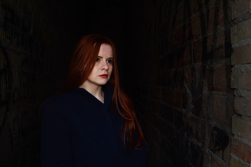 Fashion portrait of a young red-haired girl in a blue coat between the walls in the dark.