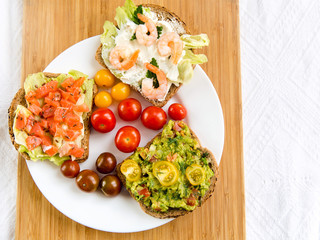Open sandviches with guacamole, fish and prawn and tomato cherry on the plate, wooden background