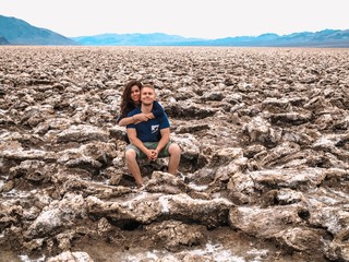 A girl with long hair hugs the neck of a blond man, he sits on salt rocks on the Devil's Golf course in the Valley of death