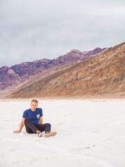 Fototapeta na wymiar The young blond man in a blue shirt and shorts is sitting on a saline soil Badwater in Death Valley