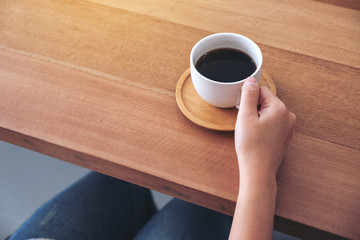 Closeup image of hand holding a white cup of hot coffee to drink on wooden table