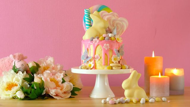 On trend Easter candy land drip cake decorated with lollipops, cand eggs and white chocolate bunny in pink party table setting.