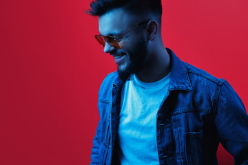 Cheerful stylish hipster bearded young man wearing yellow sunglasses and blue jeans jacket posing in neon light over red background.