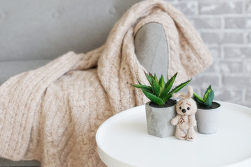 A beige plaid of handmade is lying on a gray sofa. Before the sofa stands a tabouret, on it there is a bunny of the same color as a plaid. All this on the background of a brick wall gray colour.