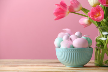 Bowl with Easter eggs and flowers on table against color background