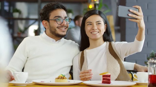 Beautiful Asian girlfriend and middle eastern boyfriend smiling and posing for smartphone camera while taking a selfie together in cafe