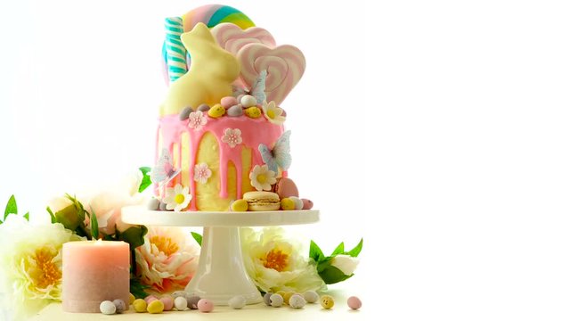 On-trend Easter theme candy land drip cake decorated with lollipops, cand eggs and white chocolate bunny on white background.
