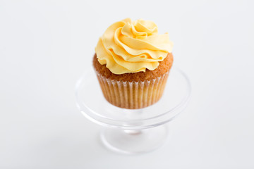 food, pastry and sweets concept - cupcake with yellow buttercream frosting on glass confectionery stand over white background