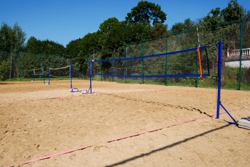 Volleyball nets on blue poles on empty sand court surrounded by lush green trees on summer day