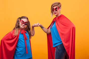 Mother and daughter dressed like heroins fist bumping and looking at the camera over yellow background.
