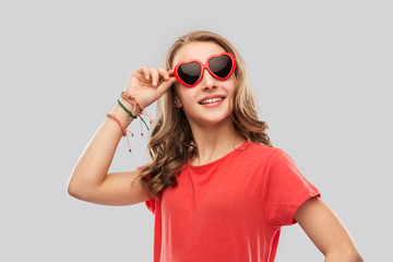 valentine's day, love and people concept - smiling pretty teenage girl wearing bangles on hand in red heart shaped sunglasses over grey background
