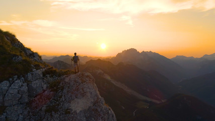 DRONE: Flying around a male hiker standing on the mountain peak at sunset.