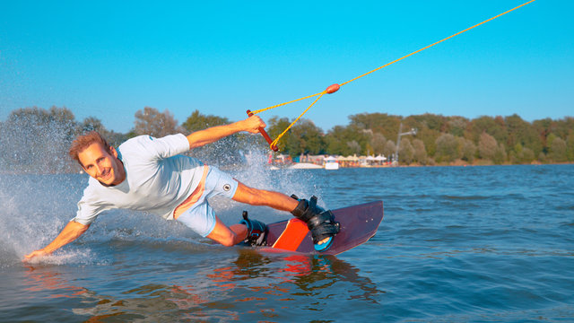 CLOSE UP: Cheerful man wakeboarding and splashing the lake water with his hand.