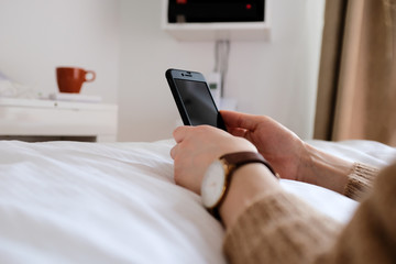 Frau sitzt auf ihrem Bett in der Wohnung und hat ein Handy in der Hand