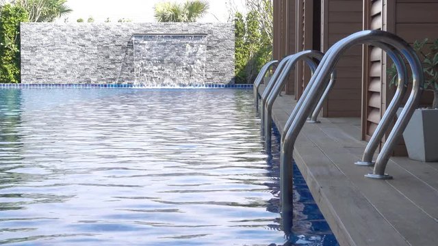 Swimming pool with metal stairs and wooden deck. Waterfall feature in the background