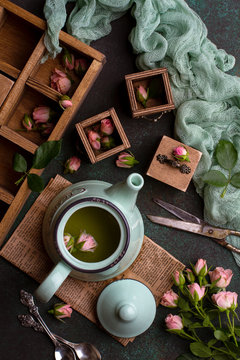 Overhead shot of composition with green teapot with tea, little wooden chest of drawers, pink roses, green gauze and old scissors on dark green concrete background