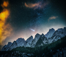 mountain range and forest with a beautiful night sky, milky way and a falling star with a man on the top of a mountain in the distance. nightscape mountains landscape in austria