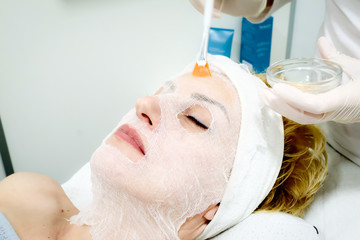 beautician worker holding paintbrush  applying facial cosmetic serum  in biological treatment to female client  in beauty salon, eyes closed, close up 