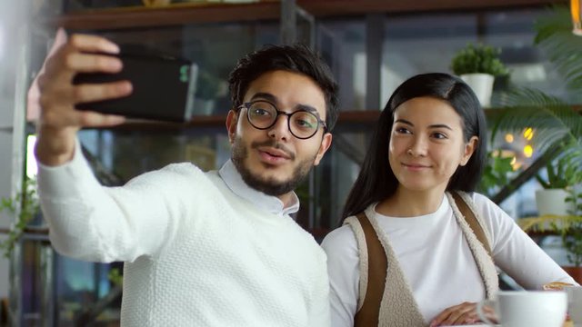 Young asian woman and middle eastern man in eyeglasses smiling and posing for smartphone camera while taking a selfie in cafe