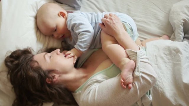 Happy young mother playing with baby in bed. The kid touches his finger to the mouth of the mother, the development of kids