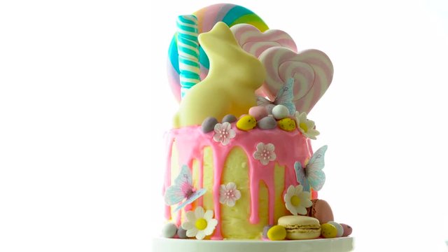 On-trend Easter theme candy land drip cake decorated with lollipops, cand eggs and white chocolate bunny on white background.