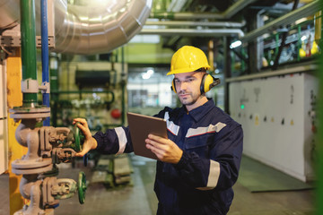 Young Caucasian hardworking heavy industry worker in protective suit and helmet using tablet and screwing valve.