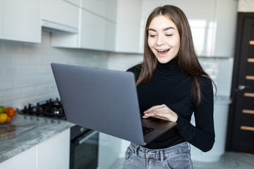 Happy young woman using laptop at counter at home in the kitchen